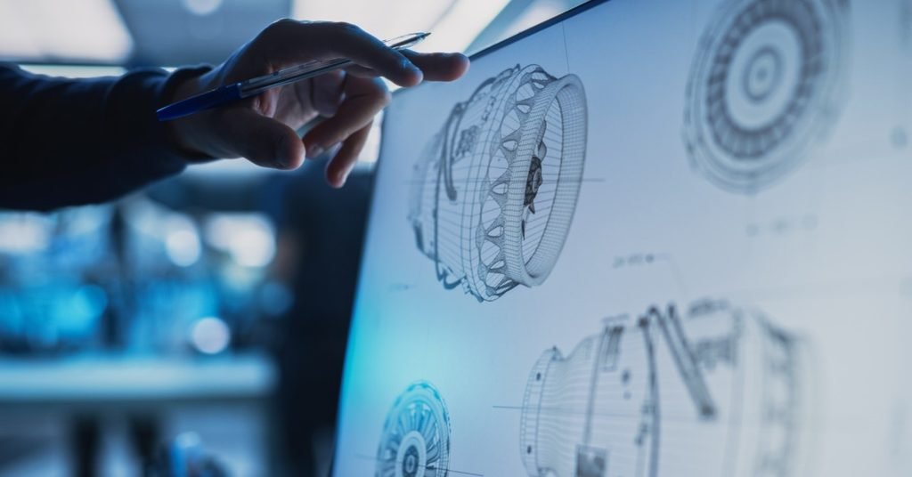 A hand holding a pen points at a monitor showing technical diagrams of circular aerospace mechanical parts in a workspace.