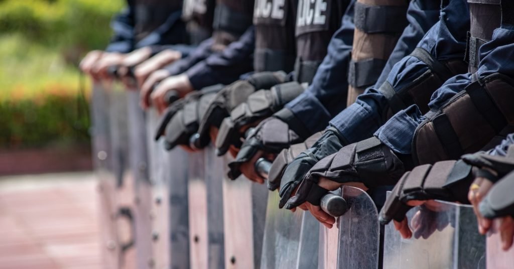 A line of people wearing police uniforms is standing outside and holding shields that are touching the ground.