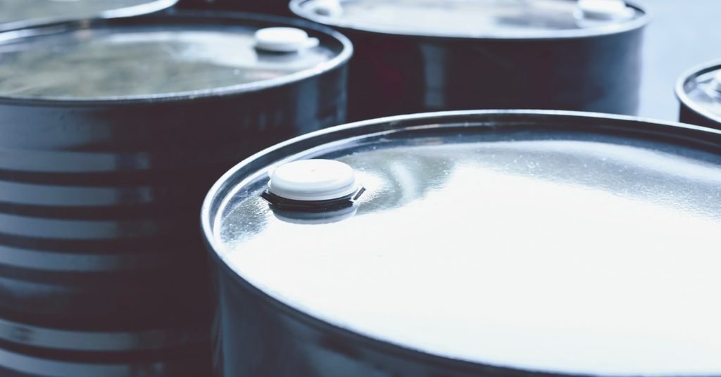 Two rows of three black metal barrels sit in a line in a warehouse. They have white caps on top.